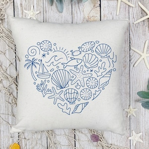 May include: A square, off-white pillow with a blue heart-shaped design filled with nautical elements. The design includes seashells, starfish, a sailboat, and a sun. The pillow is on a wooden surface with starfish and greenery.