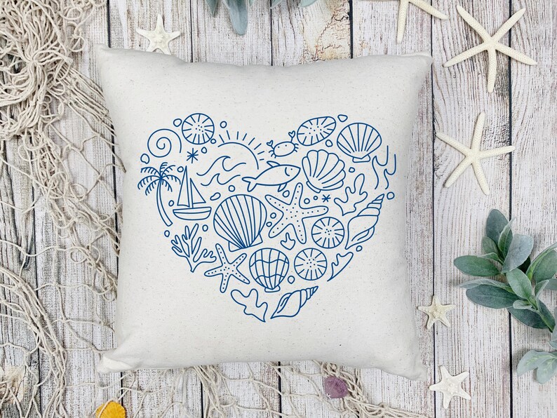 May include: A square, off-white pillow with a blue heart-shaped design filled with nautical elements. The design includes seashells, starfish, a sailboat, and a sun. The pillow is on a wooden surface with starfish and greenery.