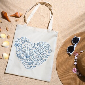 May include: A cream-colored tote bag with a blue heart-shaped design of seashells, a sailboat, and other nautical elements. The bag is on sand, with a straw hat, sunglasses, and seashells scattered around.