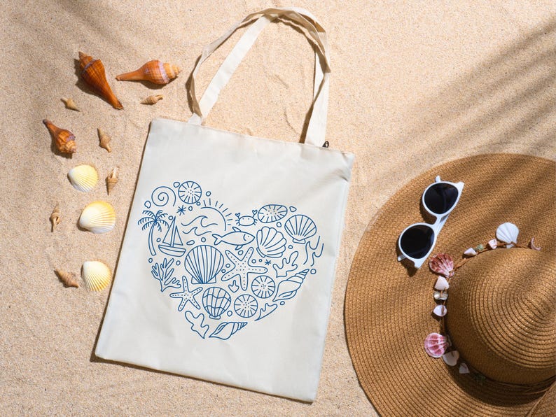 May include: A cream-colored tote bag with a blue heart-shaped design of seashells, a sailboat, and other nautical elements. The bag is on sand, with a straw hat, sunglasses, and seashells scattered around.
