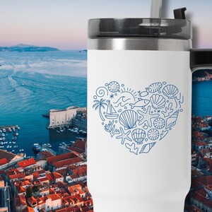 May include: White travel mug with a black handle and lid. A blue line drawing of a heart filled with seashells, a sailboat, and other nautical elements decorates the mug. The background shows a coastal city.