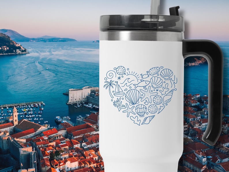 May include: White travel mug with a black handle and lid. A blue line drawing of a heart filled with seashells, a sailboat, and other nautical elements decorates the mug. The background shows a coastal city.