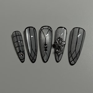 May include: Five silver-coloured, almond-shaped artificial nails. Each nail features a unique design with black lines, rhinestones, and a silver cross. The nails are arranged on a light grey surface.