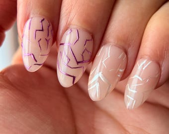 May include: Close-up of manicured nails with a nude base and abstract designs. Two nails feature a cracked, purple foil design, while the others have a silver geometric pattern. The nails are oval-shaped.