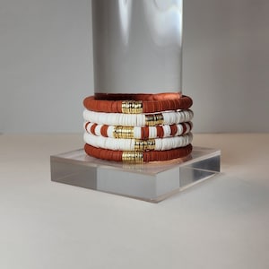 May include: Stack of five bracelets in shades of white and terracotta, with gold-colored accents. The bracelets are made of flat, disc-shaped beads. The bracelets are stacked on a clear, rectangular stand.