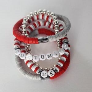 May include: A stack of four bracelets with red and white beads and silver accents. The bracelets spell out "CUSTOMIZE ME".