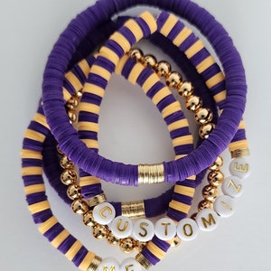 May include: A set of five bracelets with purple and yellow striped bands and gold beads. The bracelets are stacked on top of each other and spell out "CUSTOMIZE ME" in white letters with gold outlines.
