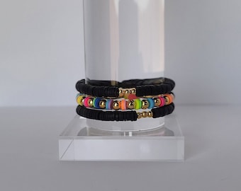 May include: Three stacked bracelets with black rubber beads and gold accents. The top bracelet has rainbow colored beads, the middle bracelet has neon pink and green beads, and the bottom bracelet has black rubber beads.