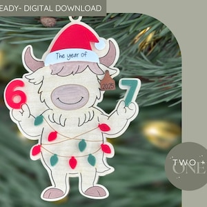 May include: Wooden Christmas ornament shaped like a cartoon yak wearing a Santa hat. The yak holds the numbers 6 and 7, with a tag reading "2025." The ornament is decorated with red and green lights and the text "The year of".