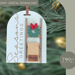 Può includere: Ornamento natalizio in legno azzurro con le parole "Season's Greetings" e un design a scala. La scala ha una ghirlanda verde con un fiocco rosso e un piccolo cartello rettangolare con "The Williams Family".