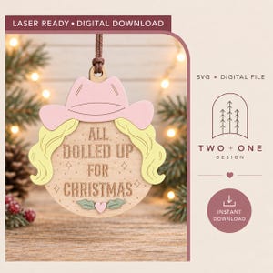 Peut inclure: Une décoration de Noël circulaire en bois avec un chapeau de cow-boy rose clair, des cheveux blonds et le texte gravé ALL DOLLED UP FOR CHRISTMAS. Le motif inclut de petites étoiles, un graphique de houx vert et un petit cœur rose en bas.