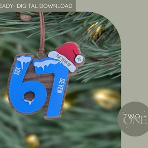 May include: Wooden Christmas ornament shaped as the number 67, with blue detailing and a Santa hat. The ornament has the text "The Year of '25" and is suspended by a brown string. The background is a blurred image of a Christmas tree.