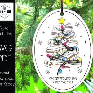 Puede incluir: Un adorno de madera ovalado blanco con una cuerda marrón para colgar. El adorno presenta un árbol de Navidad hecho de Crocs apilados en marrón y blanco con luces de colores. El texto "Crocin' Around The Christmas Tree" está impreso debajo del árbol.