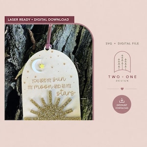 Pode incluir: Um ornamento de madeira em arco com um sol de glitter dourado na parte inferior e uma lua crescente branca com uma estrela amarela no topo. O texto diz "you are my sun my moon and all my stars", rodeado por pequenas estrelas gravadas e um cordão rosa.