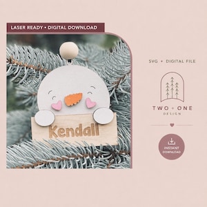 May include: A laser-ready digital download file featuring a snowman ornament design. The snowman has a light-colored head, a small orange carrot nose, two pink heart-shaped cheeks, and a wooden sign below with the name Kendall engraved.