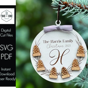 May include: White round bauble with a wreath and pine cones. The text "The Harris Family Christmas 2023" is written on the bauble. The letter "H" is in the centre of the bauble. Each pine cone has a name tag with a name on it: Ashton, Owen, Kevin, Cindy, Kenzie, and Teddy.
