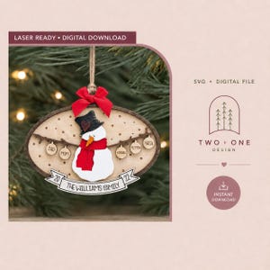 Könnte beinhalten: Ein ovaler Weihnachtsbaumschmuck aus Holz mit einem weißen Schneemann, schwarzem Hut, oranger Nase und rotem Schal. Ein rotes Band hängt oben. Ein Banner unten zeigt 2022 und THE WILLIAMS FAMILY, mit kleinen runden Anhängern beschriftet mit DAD, MOM, KENDALL, PEYTON und NASH.
