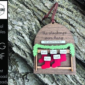 Op de afbeelding: Houten kerstornament met de tekst "The stockings were hung" en "The Williams Family". Het ornament heeft rode kousen met namen, een groene slinger en een bruine leren ophanglus. Het ontwerp is laser-klaar.