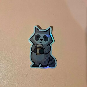 May include: A sticker of a gray raccoon holding a brown and white coffee cup. The raccoon has a black nose and black eyes. The sticker has a holographic finish.