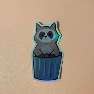 May include: A sticker of a cartoon raccoon sitting in a blue trash can. The raccoon is gray and has a black nose and eyes. The trash can has a blue and white striped pattern.