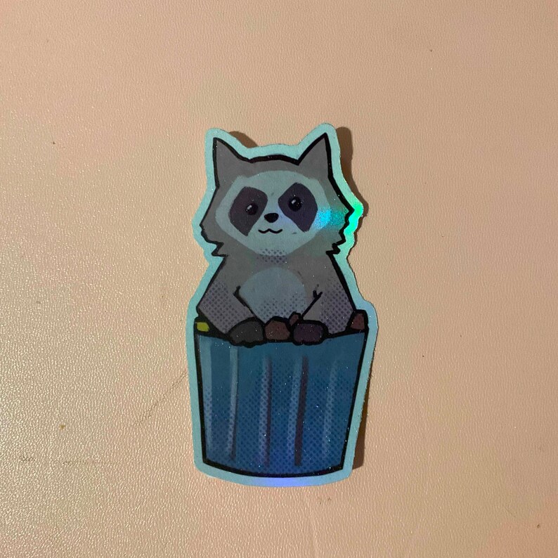 May include: A sticker of a cartoon raccoon sitting in a blue trash can. The raccoon is gray and has a black nose and eyes. The trash can has a blue and white striped pattern.