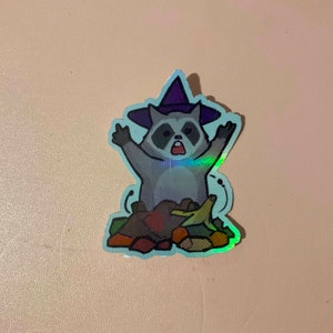 May include: A holographic sticker of a raccoon wearing a witch hat and holding a bunch of leaves. The raccoon has a surprised expression on its face.