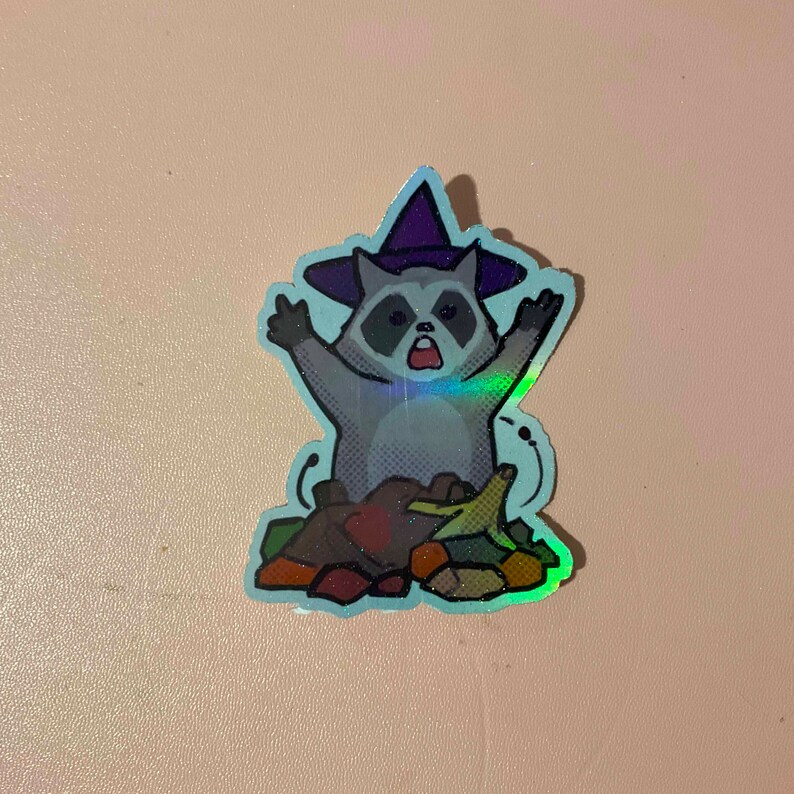May include: A holographic sticker of a raccoon wearing a witch hat and holding a bunch of leaves. The raccoon has a surprised expression on its face.