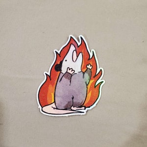 May include: A sticker featuring a cartoon opossum with a surprised expression, standing in a bright orange and yellow flame. The opossum is wearing a grey, textured, hooded garment.
