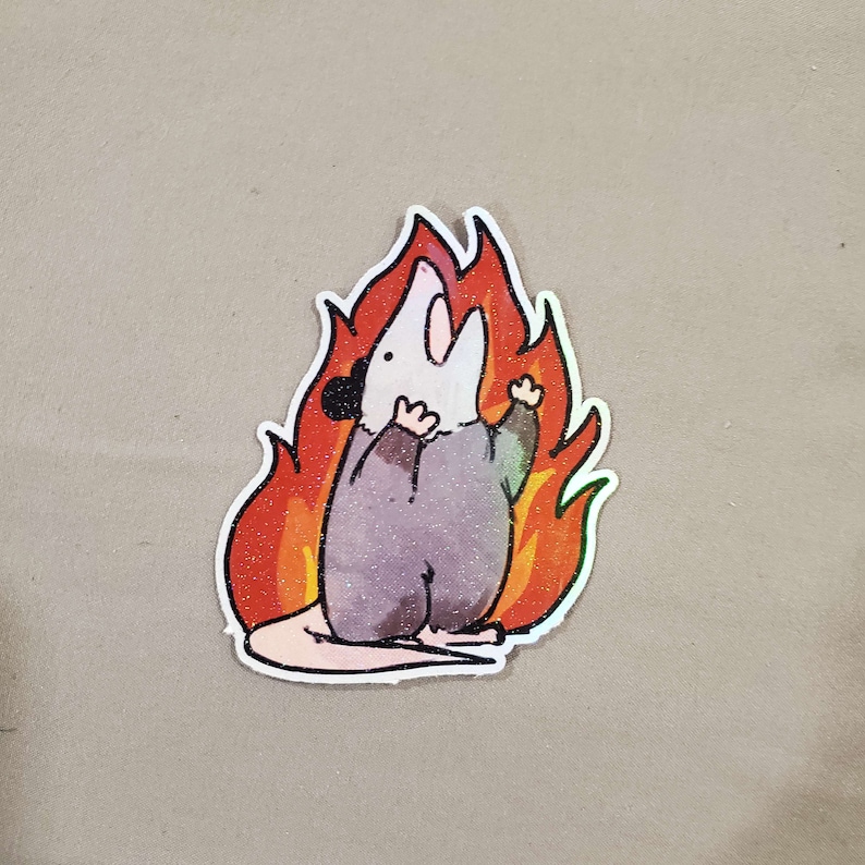 May include: A sticker featuring a cartoon opossum with a surprised expression, standing in a bright orange and yellow flame. The opossum is wearing a grey, textured, hooded garment.