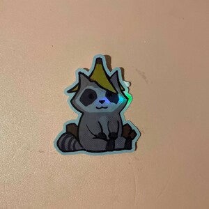May include: A sticker of a cartoon raccoon wearing a banana on its head. The raccoon is sitting down with its paws in front of it. The sticker has a holographic finish.