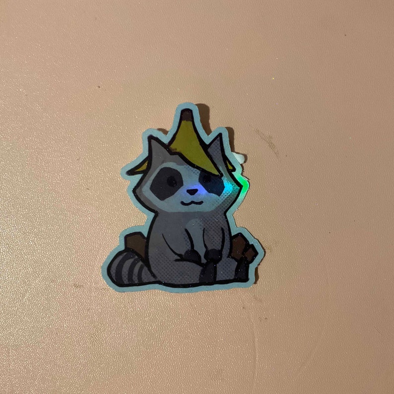 May include: A sticker of a cartoon raccoon wearing a banana on its head. The raccoon is sitting down with its paws in front of it. The sticker has a holographic finish.