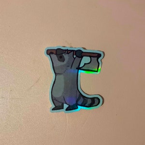 May include: A holographic sticker of a raccoon holding an axe above its head. The raccoon is gray with a black nose and eyes. The axe is brown with a silver blade.