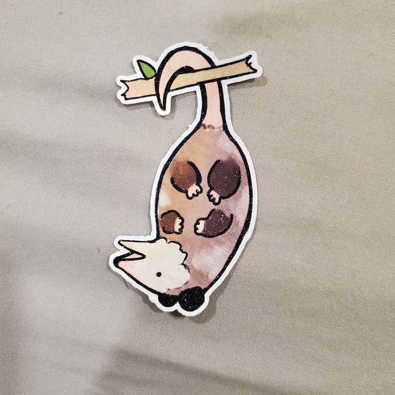 May include: A brown and white possum with a pink tail hanging upside down from a branch. The possum has a white face and black eyes. The branch is brown and has a green leaf on it.