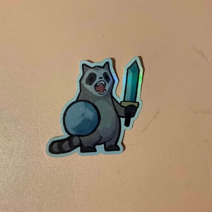 May include: A sticker of a cartoon raccoon holding a sword. The raccoon is wearing a blue and white shield. The sticker is holographic.