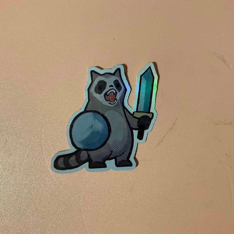 May include: A sticker of a cartoon raccoon holding a sword. The raccoon is wearing a blue and white shield. The sticker is holographic.