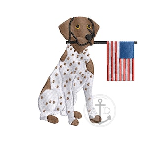 以下が含まれることがあります： アメリカ国旗を口にくわえた犬の刺繍デザイン。犬は白地に茶色の斑点と茶色の頭をしています。旗は赤と白のストライプと白い星の青い正方形を持っています。