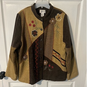 以下が含まれることがあります： 赤、茶色、緑の色合いの刺繍された花が施された、茶色とベージュのパッチワークジャケット。ジャケットはボタン留めと立て襟です。ラベルには「Graff」と書いてあります。