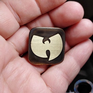 Puede incluir: Un pin de latón de forma cuadrada con un logotipo dorado de Wu-Tang Clan. El logotipo es una 'W' estilizada con un círculo alrededor.