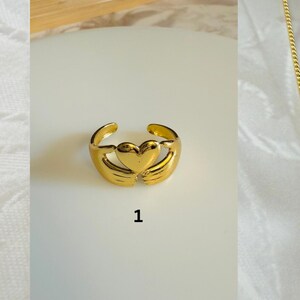 May include: Gold-colored ring with a heart-shaped design. The ring features a heart in the center, embraced by two curved arms. The number "1" is printed below the ring. The ring is on a white surface.