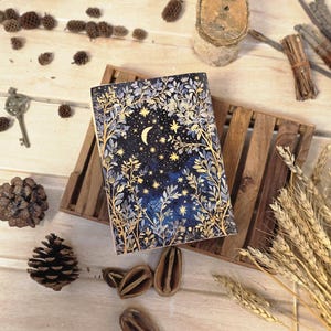 Puede incluir: Un diario de tapa dura con un dise&ntilde;o celestial. La portada presenta un fondo azul oscuro con una luna creciente, estrellas e ilustraciones bot&aacute;nicas doradas y blancas. El diario est&aacute; sobre una bandeja de madera, rodeado de elementos decorativos.