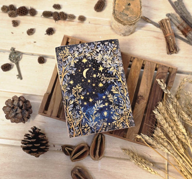 Puede incluir: Un diario de tapa dura con un dise&ntilde;o celestial. La portada presenta un fondo azul oscuro con una luna creciente, estrellas e ilustraciones bot&aacute;nicas doradas y blancas. El diario est&aacute; sobre una bandeja de madera, rodeado de elementos decorativos.