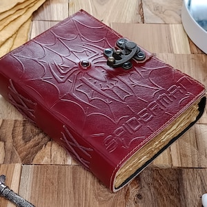 May include: A burgundy leather journal with a spiderweb and spider design embossed on the cover. The word "SPIDERMAN" is embossed on the bottom right corner. The journal has a metal clasp closure.