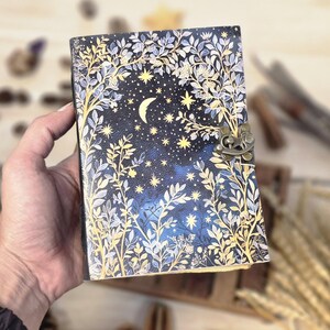 Puede incluir: Un diario decorativo con un dise&ntilde;o celestial. La portada presenta un fondo azul oscuro con una luna creciente, estrellas y detalles florales en dorado y blanco. Tiene un cierre de metal.
