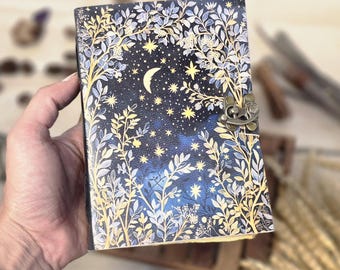May include: A decorative journal with a celestial design. The cover features a dark blue background with a crescent moon, stars, and floral accents in gold and white. It has a metal clasp closure.