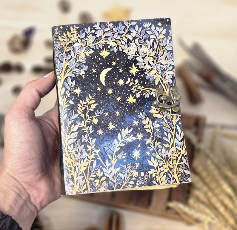 Puede incluir: Un diario decorativo con un dise&ntilde;o celestial. La portada presenta un fondo azul oscuro con una luna creciente, estrellas y detalles florales en dorado y blanco. Tiene un cierre de metal.