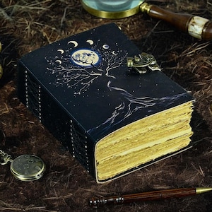 Könnte beinhalten: Ein schwarzes Notizbuch mit einem Baum- und Monddesign auf dem Einband. Das Notizbuch hat einen Metallverschluss und goldfarbene Seiten. Eine Vintage-Taschenuhr und ein Stift sind ebenfalls sichtbar.