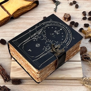 May include: A black leather journal with an embossed tree design and a metal clasp. The pages have a vintage, aged appearance. The journal is closed with a black leather strap and sits on a wooden surface. A key and other decorative items are in the background.