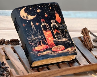 Op de afbeelding: Een zwart leren dagboek met een hemels ontwerp met een maan, sterren en illustraties van flessen en planten. Het dagboek heeft een metalen sluiting en verouderde, vergeelde pagina's, wat wijst op een vintage stijl.