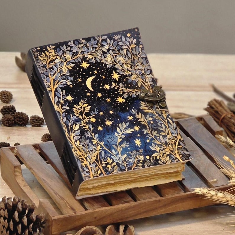 Puede incluir: Un diario decorativo con un dise&ntilde;o celestial. La portada presenta un fondo azul oscuro con una luna creciente, estrellas y follaje dorado. El diario tiene un cierre de metal y p&aacute;ginas envejecidas.