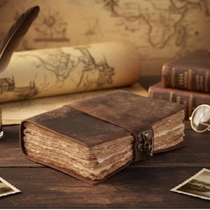 May include: A vintage-style leather journal with a clasp closure and a strap. The journal has aged, textured pages and a dark brown cover. Other items in the image include a rolled-up map, a quill, and a pair of glasses.